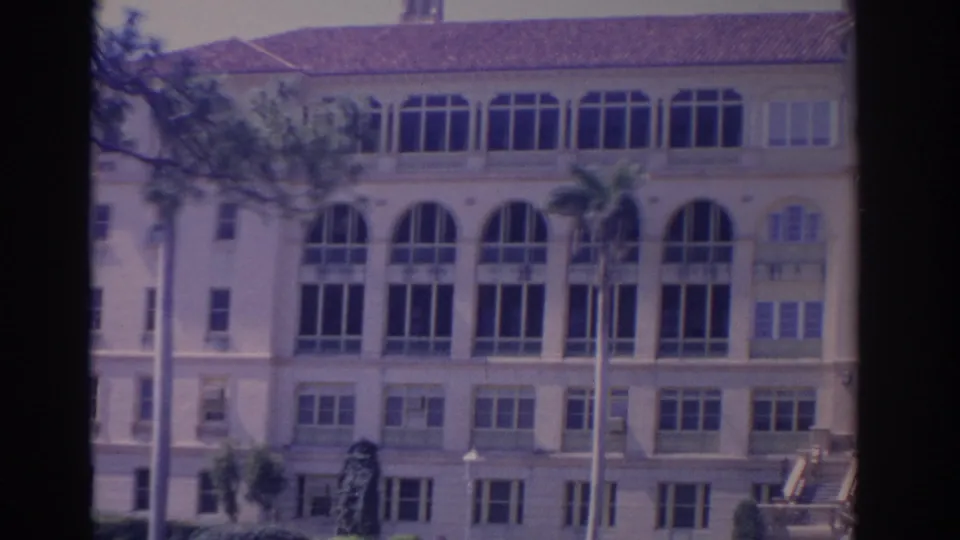 1962 Florida 8mm view of the building facade with tall arched windows, small shutters, palm trunks, and a red tile roof.