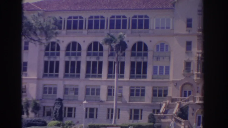 1962 Florida home-movie frame showing a closer portion of the cream building, arched windows, palms, shutters, and trimmed shrubs.