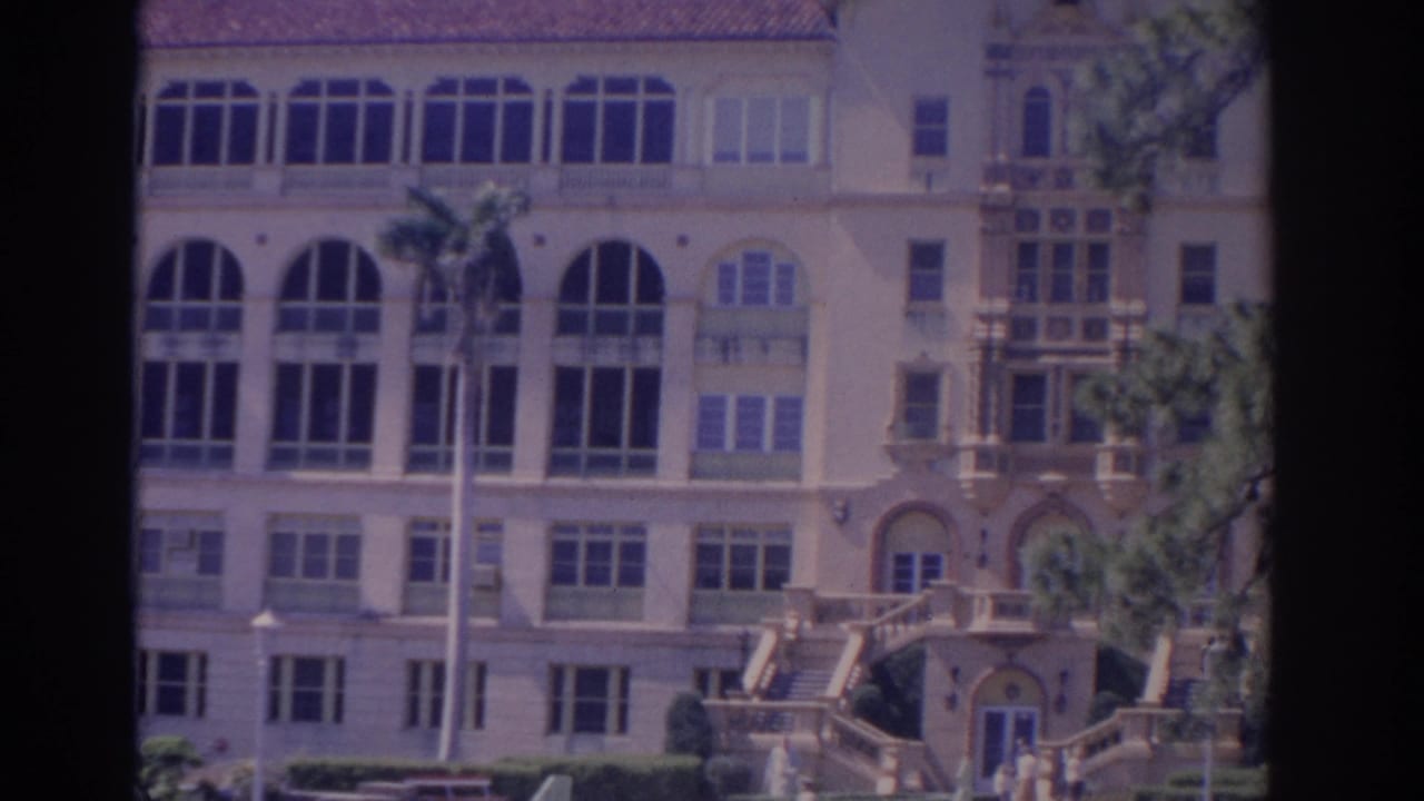 Grand cream-colored building facade with arched windows, stairway, palms, and small figures near the entrance.