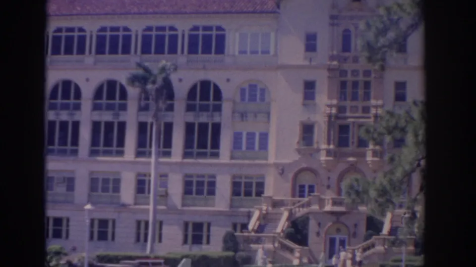 1962 Florida home-movie view of a grand cream-colored building facade with arched windows, balconies, front steps, palms, and distant visitors.