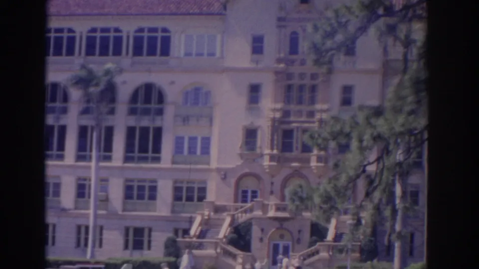 1962 Florida exterior shot showing the ornate building entrance, arched windows, stair railings, palms, hedges, and distant people at the steps.