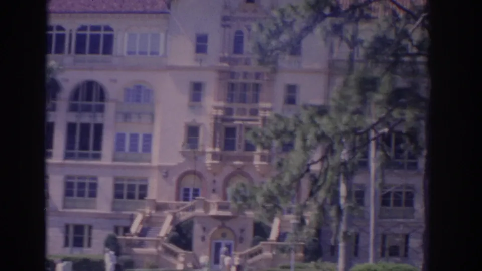 1962 Florida home-movie frame with the building facade partly obscured by trees, showing arched windows, balconies, front stairs, and visitors.