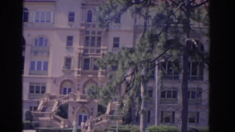 1962 Florida exterior view of the grand facade through foreground trees, with arched windows, balconies, landscaped hedges, and distant people.