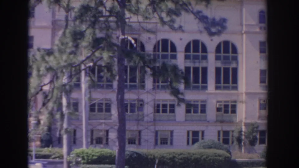 1962 Florida 8mm frame showing the lower portion of a large pale building, arched windows, shrubs, lawn, and a pine tree foreground.