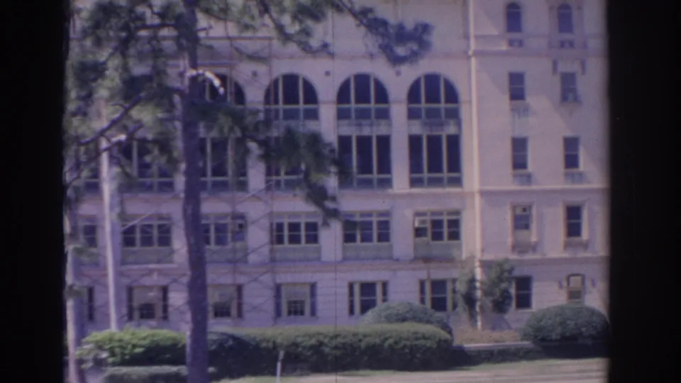 1962 Florida 8mm frame of a large light-colored building with arched windows, rows of smaller windows, shrubs, and trees in front.