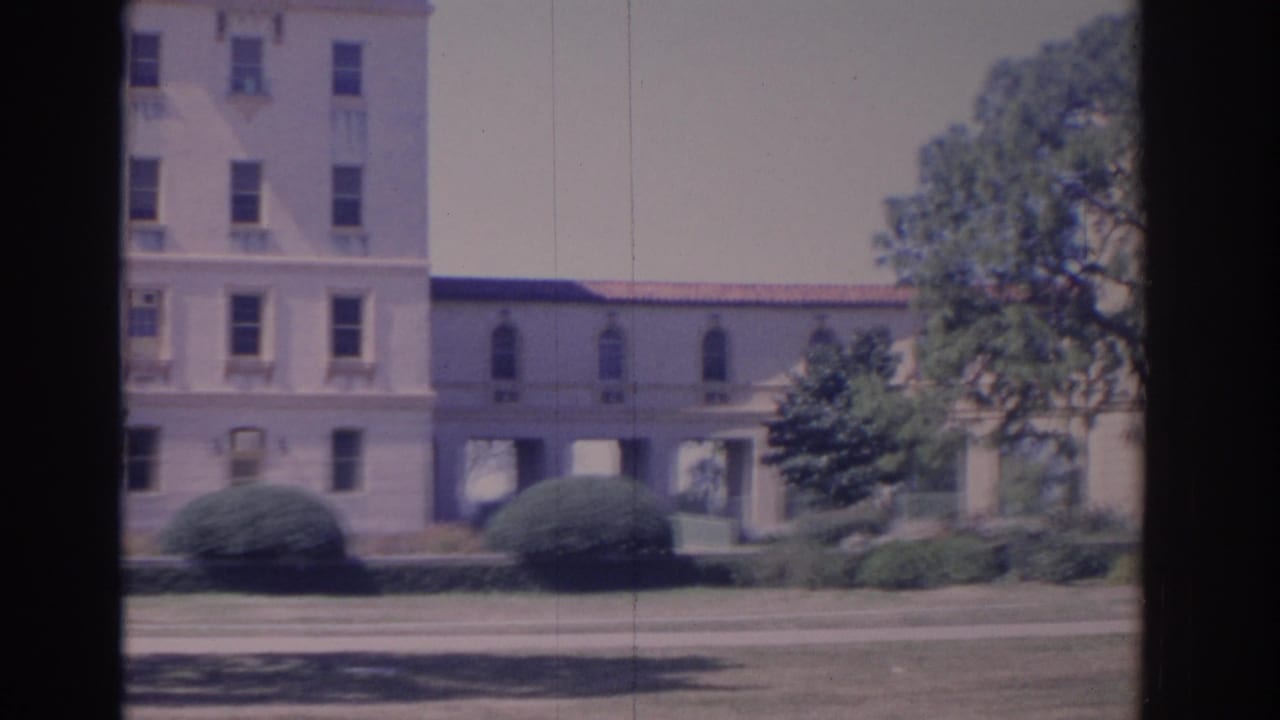 Building facade with arched windows, covered connector, shrubs, driveway, and trees.