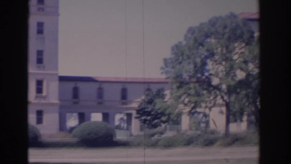 1962 Florida home-movie frame showing the side of a light institutional building with red tile roofline, covered openings, shrubs, and trees.
