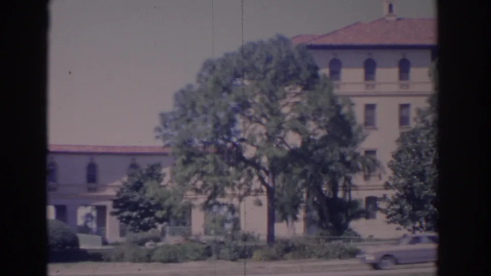 1962 Florida 8mm frame of the same cream building and mature trees, with a blurred car moving across the foreground road.