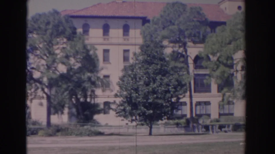 1962 Florida 8mm frame with a cream multi-story facade, arched upper windows, red tile roof, trees, lawn, and paved frontage.