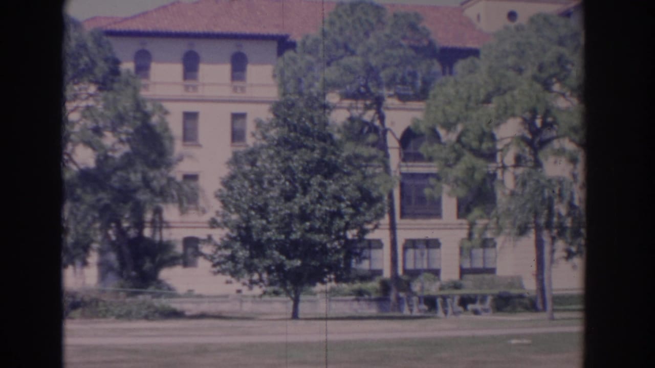 Pale multi-story building behind trees with a red tile roof and lawn in front.