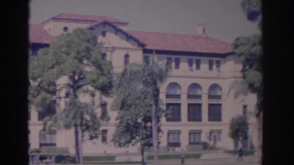 1962 Florida 8mm frame of an institutional exterior with tall arched windows, red tile roof, trees, grass, and one distant walker.