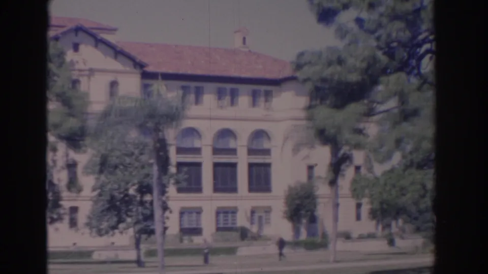 1962 Florida home-movie frame showing a tile-roofed building with arched windows, trees, lawn, bench, and distant pedestrian movement.