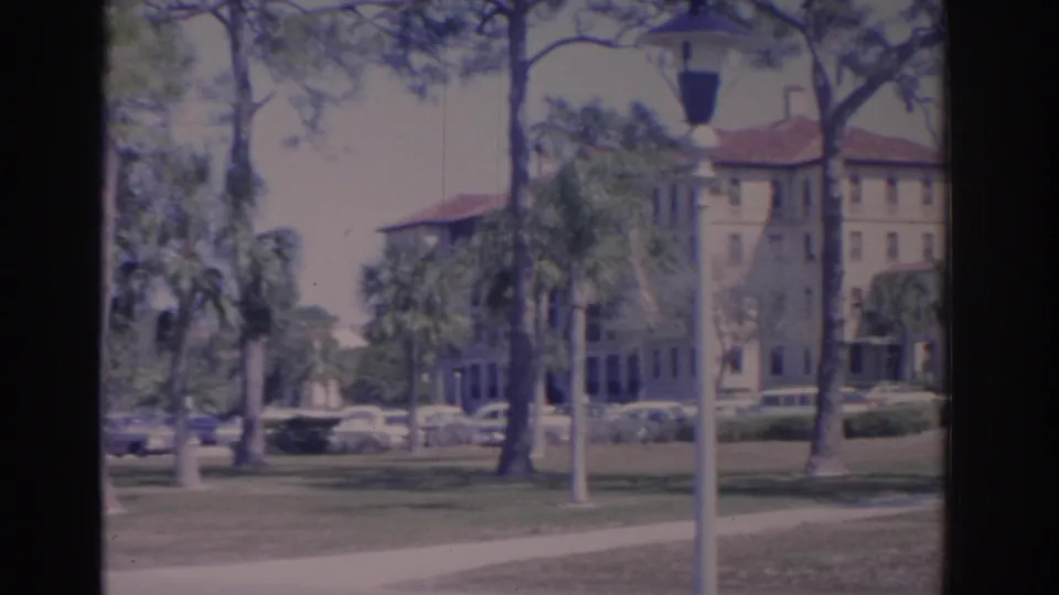 1962 Florida campus exterior with a white lamppost in front of palms, pines, lawn, parked cars, and a red-roofed building.