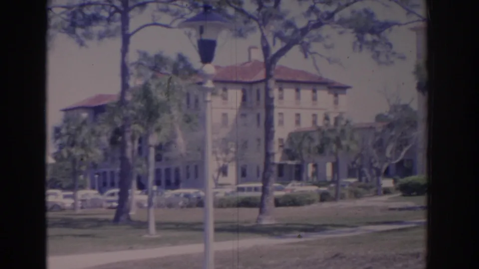 1962 Florida institutional campus building seen across a lawn, with a lamppost, palms, pines, shrubs, and parked cars.