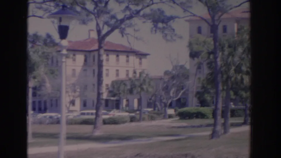 1962 Florida campus grounds with a red-roofed building, white lamppost, palms, pines, shrubs, lawn, and parked cars.
