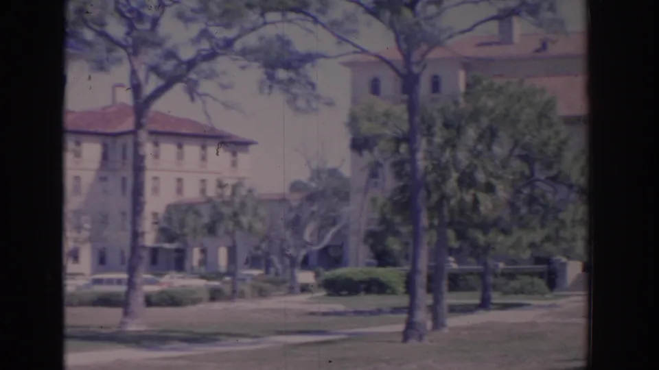 1962 Florida campus grounds with pine trees, lawn, sidewalks, and Spanish Revival-style buildings in soft 8mm color.