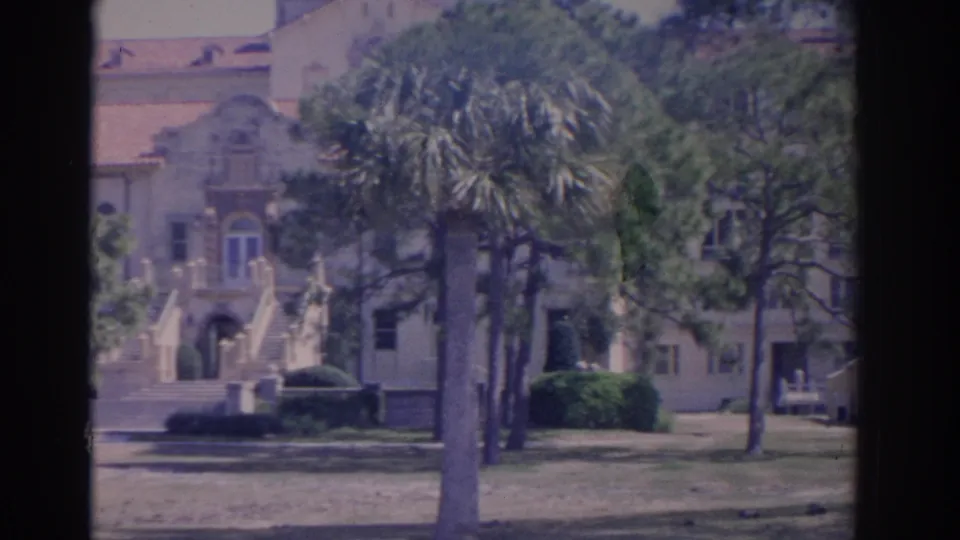 1962 Florida campus grounds with a palm tree in front of a Spanish Revival-style entrance, lawn, stairs, and a car near the right edge.