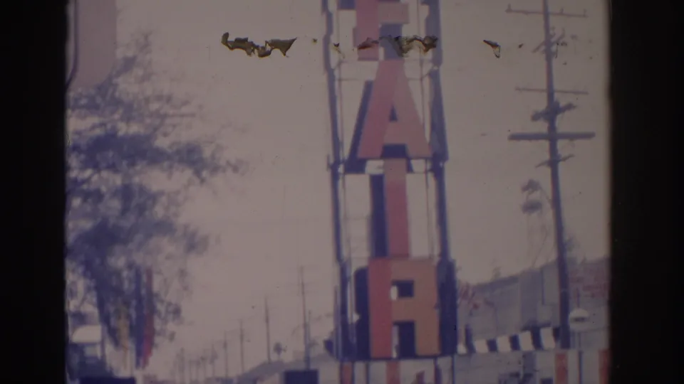 1962 Florida 8mm frame showing the FAIR sign from the sidewalk area, with trees, utility poles, and street-side structures in soft focus.