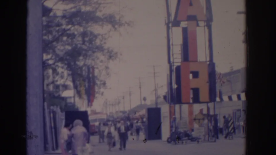 1962 Florida street frame showing the tall FAIR sign at right, pedestrians in the distance, sidewalks, poles, trees, and roadside buildings.