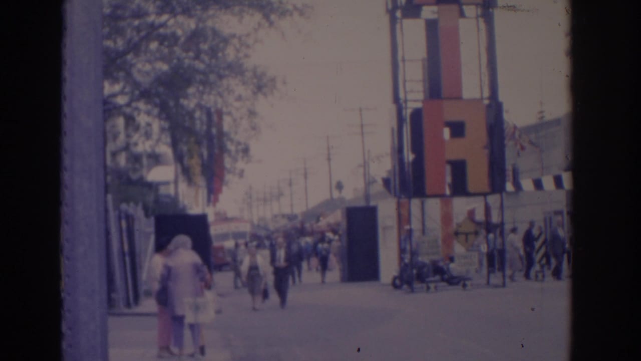 Pedestrians walk near a waterfront structure with boats and harbor activity in the distance.