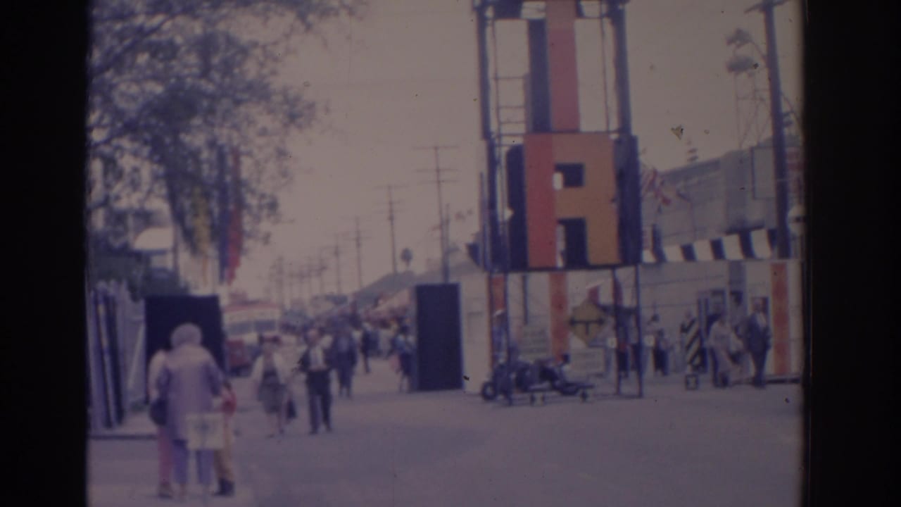 People walk along a harbor promenade with a colorful vertical structure and boats nearby.