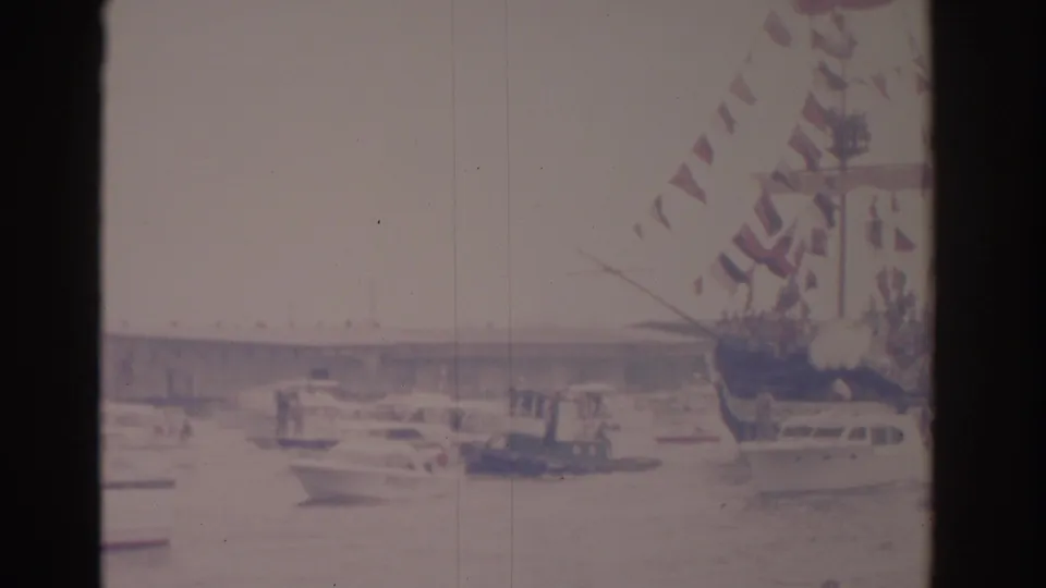 A decorated vessel with red-and-white pennants sits among small boats in a soft 1962 Florida harbor home movie frame.