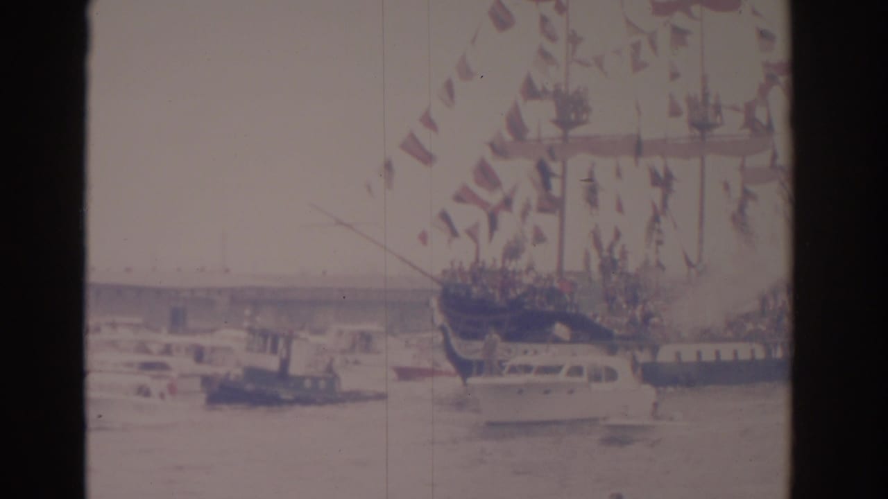 Decorated sailing ship with flags in a harbor beside smaller boats.