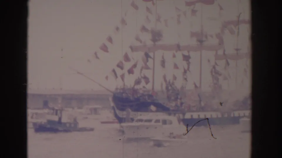 1962 Florida home-movie frame with cannon smoke near a decorated sailing ship and nearby harbor boats.