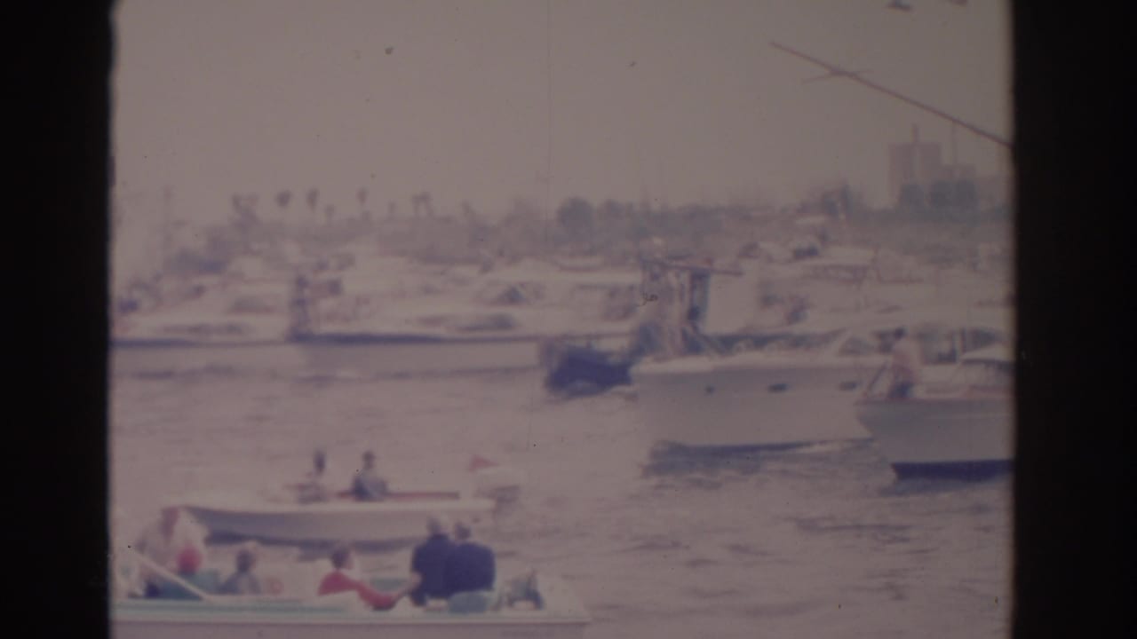 Small boats move through a marina with people aboard and larger boats in the background.