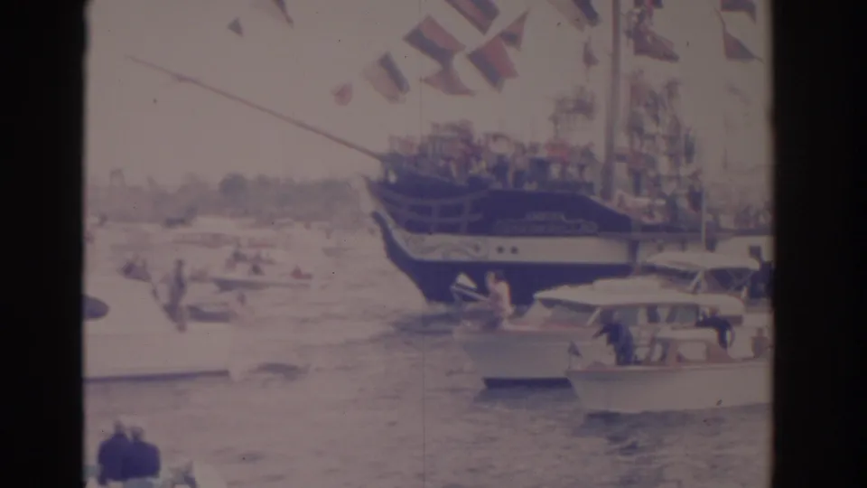 Florida 1962 home-movie frame with a flag-decorated sailing vessel, nearby motorboats, and people visible on small craft.