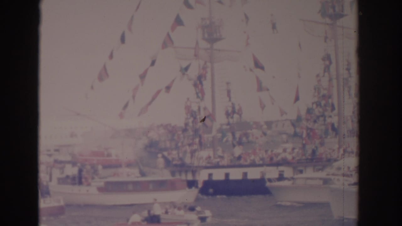 Decorated ship with flags moves through a crowded harbor with small boats nearby.