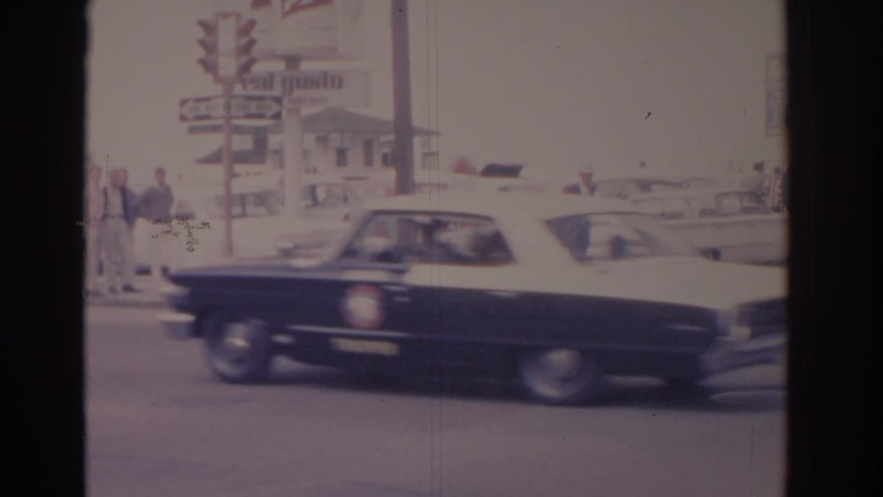 A black and white mid-century sedan moves through a Florida street scene near pedestrians, poles, and commercial roadside signage.