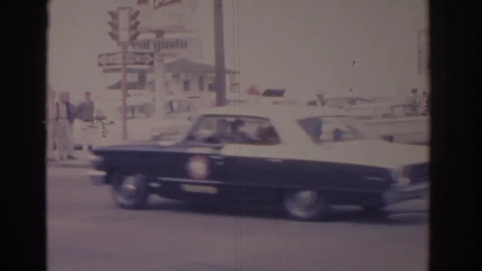 A black and white mid-century sedan moves through a Florida street scene near pedestrians, poles, and commercial roadside signage.