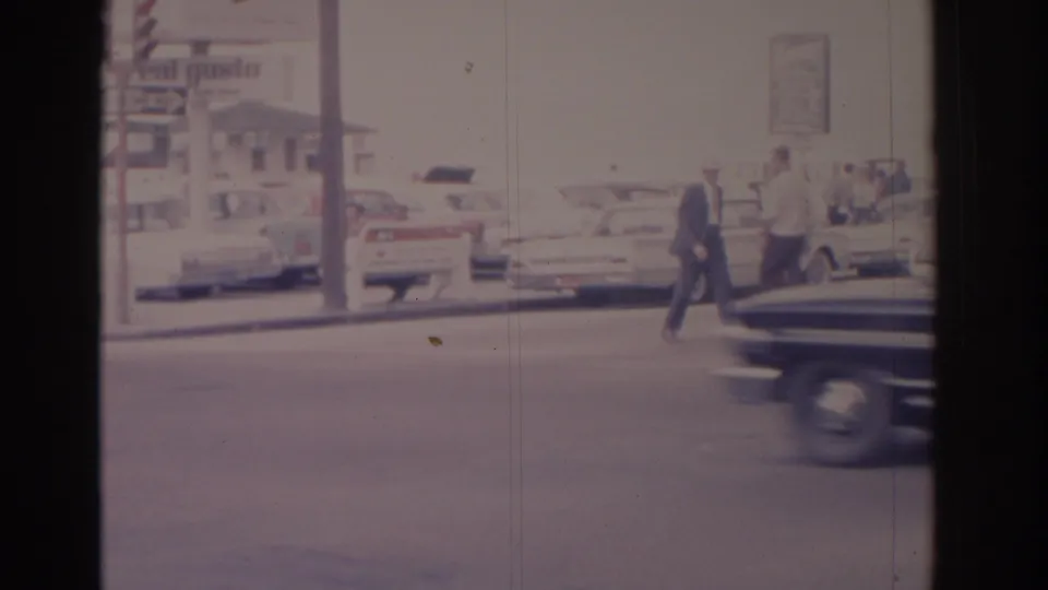 Street-level 8mm view of pedestrians in 1962 Florida crossing near parked classic cars, commercial signage, and a passing foreground vehicle.