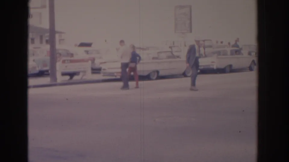 Late-frame street view from the supplied 1962 Florida footage with pedestrians, parked mid-century cars, sign poles, and a hazy roadside setting.