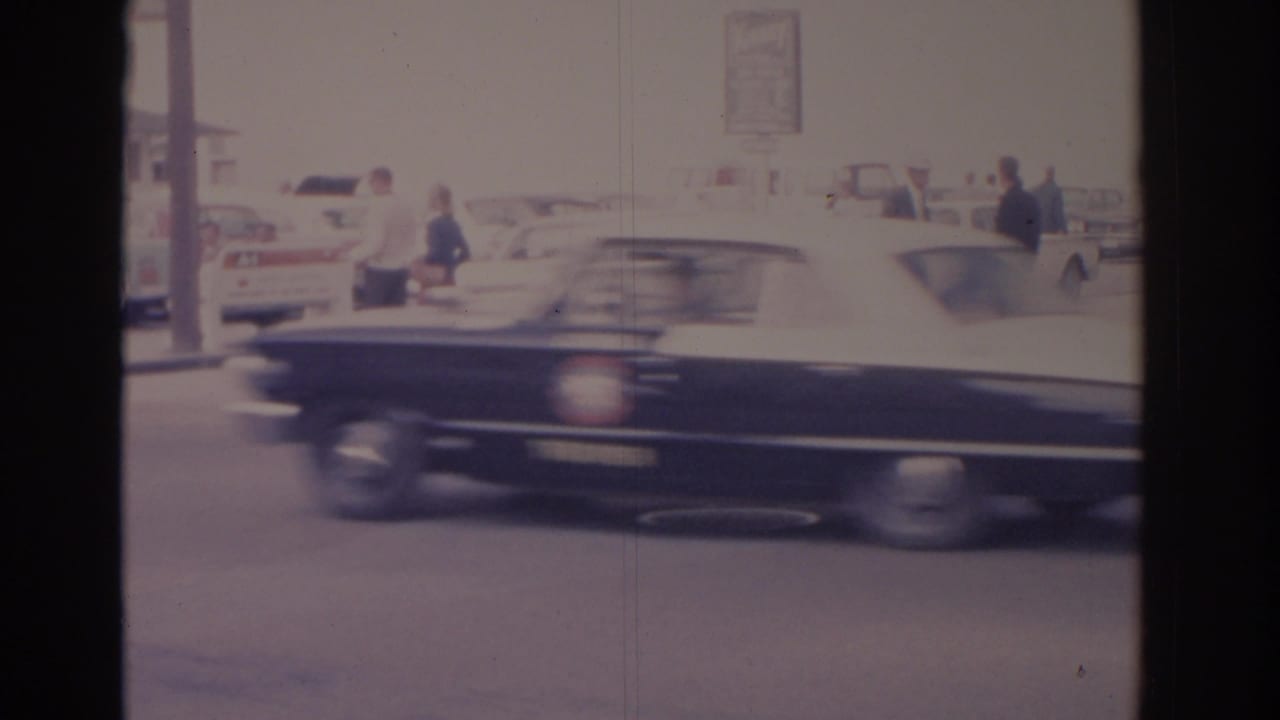 Dark car passing in front of parked classic cars and pedestrians on a bright street.