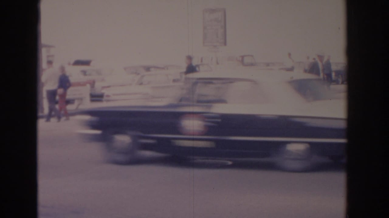 Large dark car moves past pedestrians and parked cars on a sunlit street.