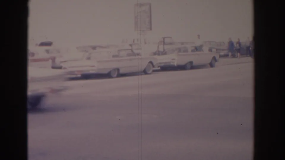 The frame settles on light-colored classic cars, pedestrians, and a roadside sign on a 1962 Florida street.