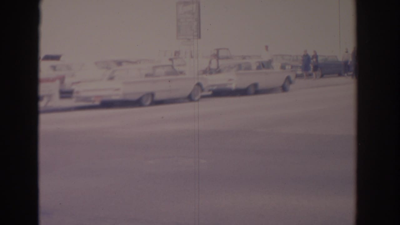 Wide city street with parked cars and distant pedestrians on a hazy 8mm frame.