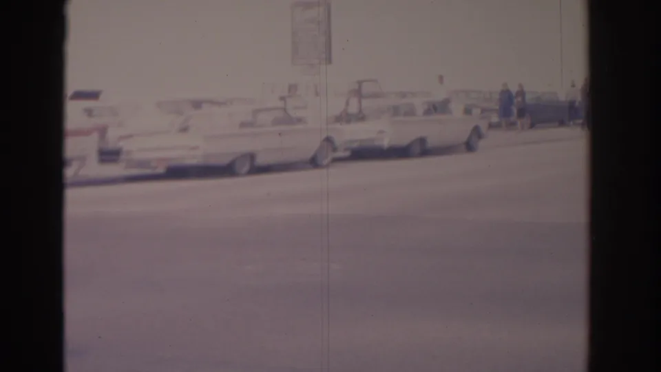 1962 Florida 8mm frame showing a wide city street, parked mid-century cars, and small figures near the curb in soft exposure.