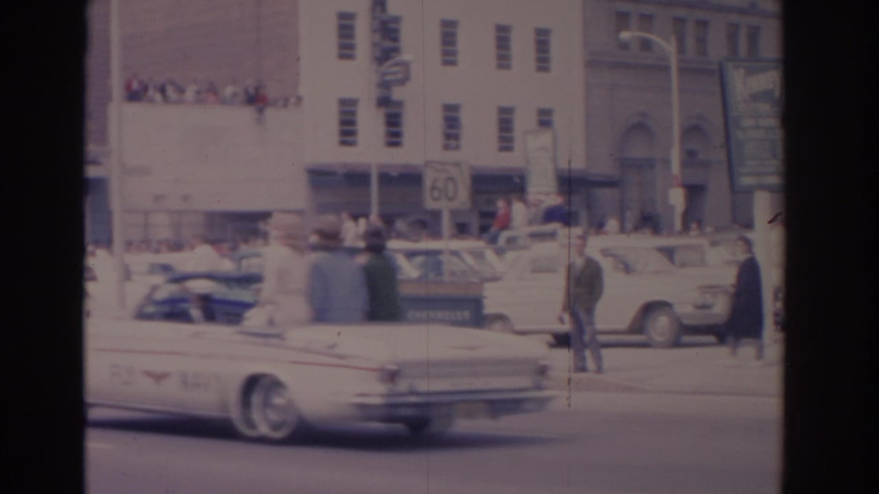 1962 Florida street scene with a white convertible moving past downtown buildings, pedestrians, rooftop onlookers, and a visible 60 sign.