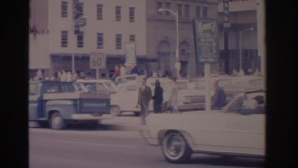 1962 Florida 8mm frame with a blue pickup, white convertible, pedestrians, storefront buildings, and rooftop spectators along a city street.