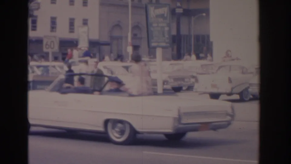 1962 Florida frame showing a white convertible marked FLY NAVY moving past pedestrians, parked cars, and downtown building signs.