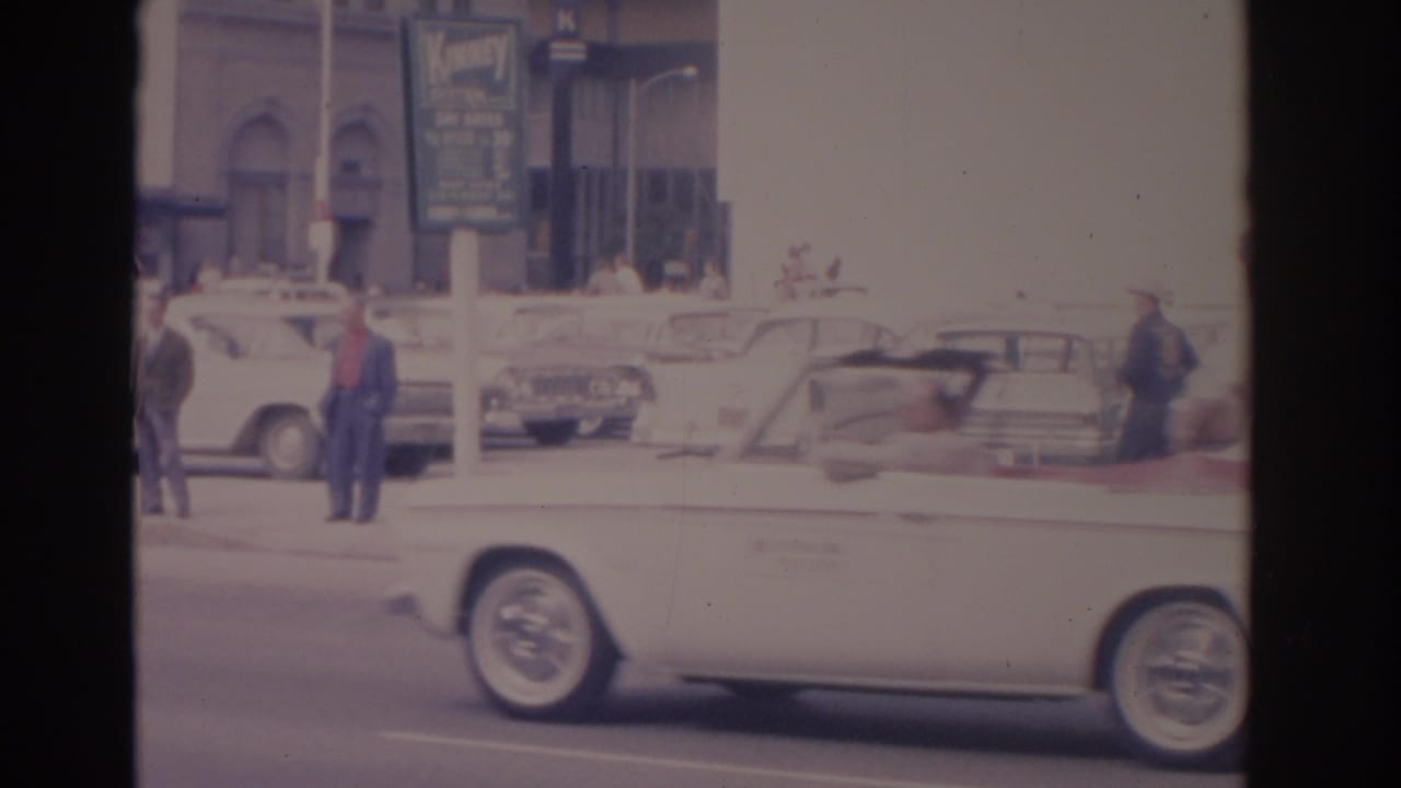 White convertible passes in front of pedestrians, parked cars, and a Kinney System sign.