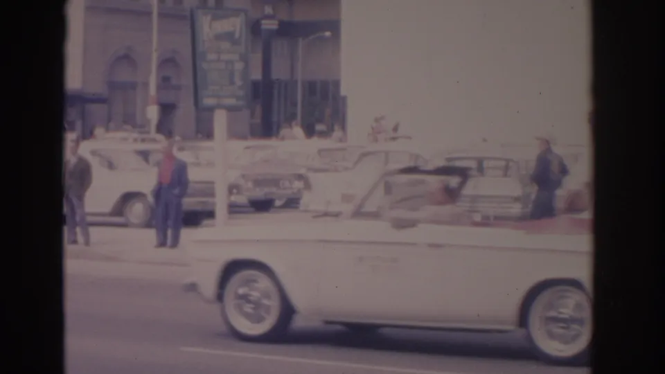 A white convertible moves through a 1962 Florida street scene with pedestrians, parked cars, and visible commercial signage.