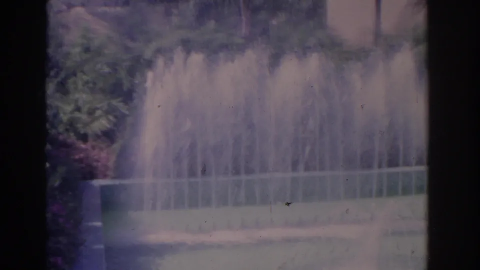 Soft color 8mm frame of a garden fountain with water jets, surrounding greenery, and flower beds.