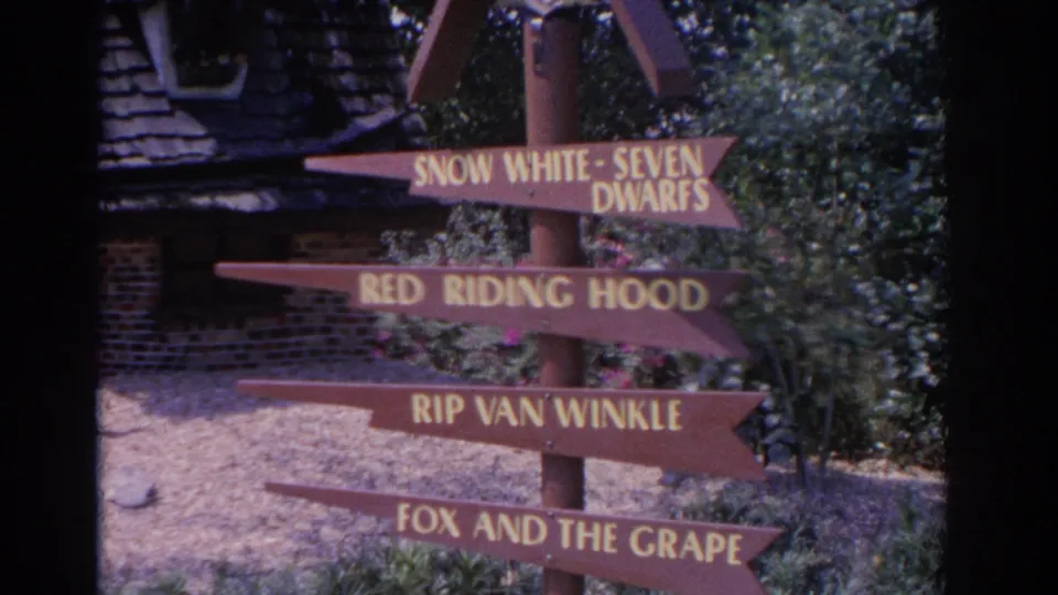 1962 Florida home-movie frame of a rustic wooden fairy-tale signpost beside shrubs and a brick building.