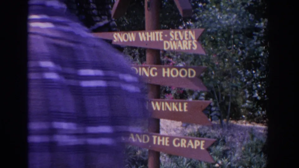 A passerby partly obscures a 1962 Florida 8mm view of a wooden signpost with fairy-tale title lettering and greenery behind it.
