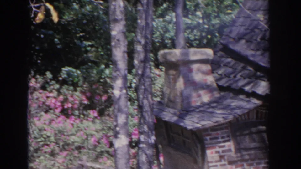 1962 Florida 8mm frame showing a miniature brick cottage with a chimney and shingled roof, surrounded by trees, foliage, and pink flowers.
