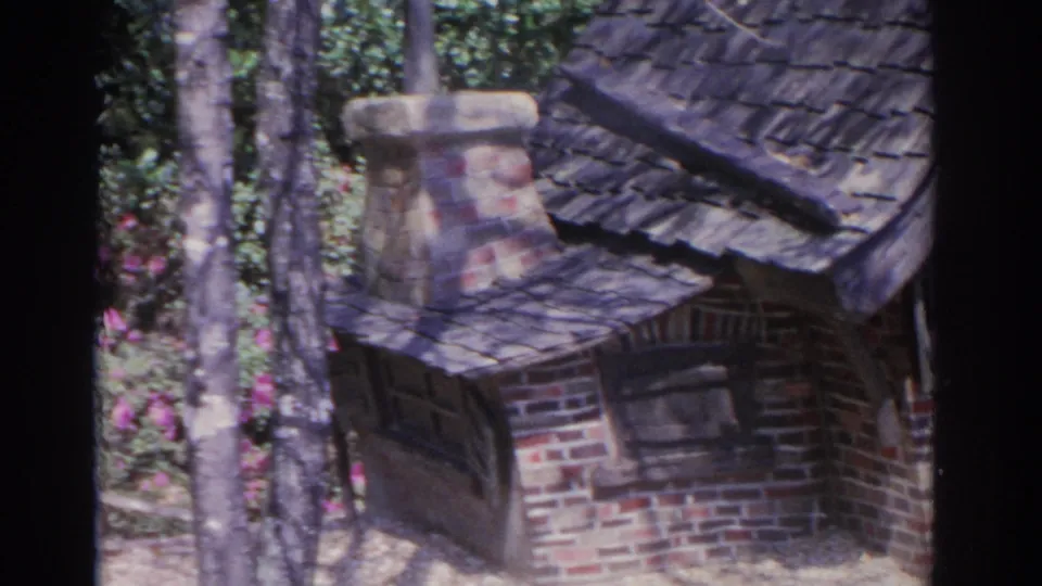 1962 Florida 8mm frame centering the miniature brick cottage, with its chimney, roof shingles, window, tree trunks, and flowering garden plants.